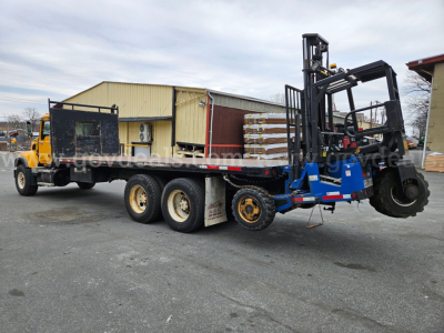 Flatbed truck with a large forklift mounted on a lowboy trailer; rear view shows three axles, yellow cab, black chassis, and forklift forks raised on blue frame.
