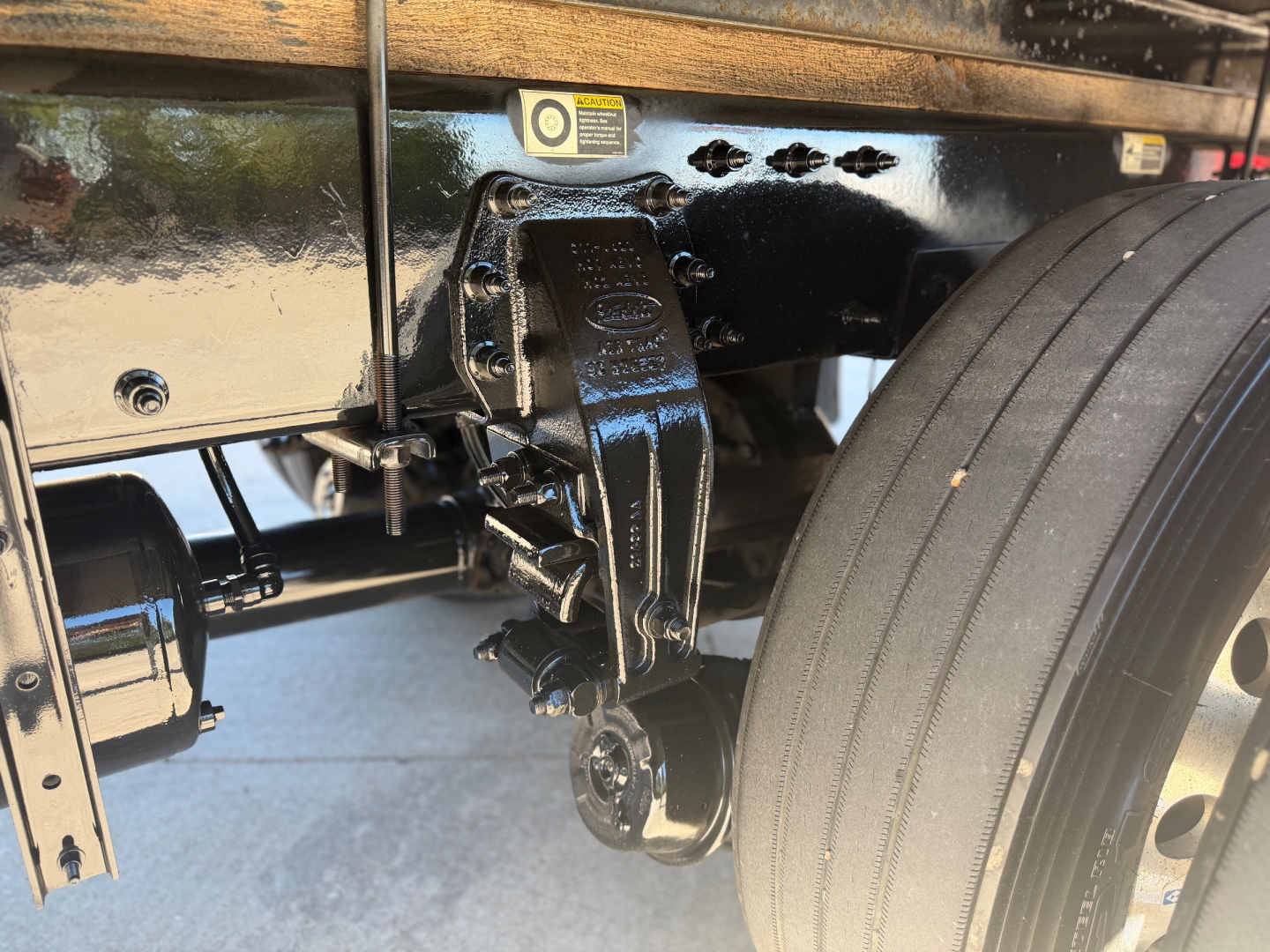 Truck frame showing front suspension with leaf spring and steering knuckle; visible riveted bracket and wheel with tire. Text on component reads 'Quality' and 'GVM' not fully legible.