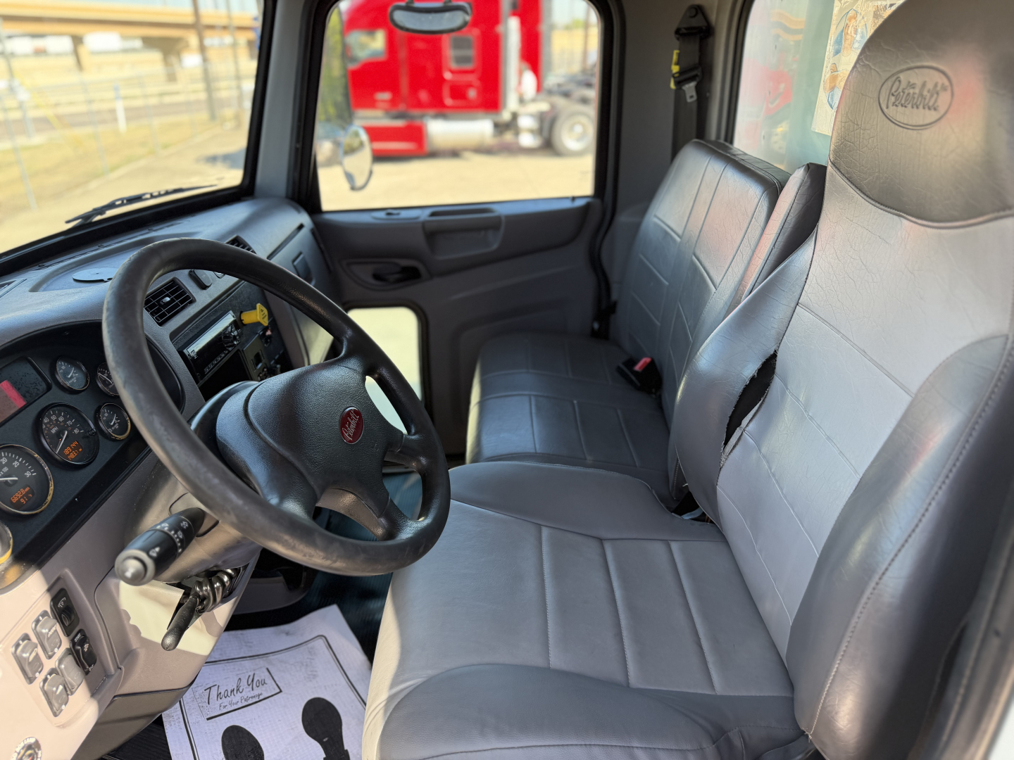 Interior of a 2020 Peterbilt 337 cab showing gray cloth seats and steering wheel with Peterbilt logo, dashboard gauges visible.