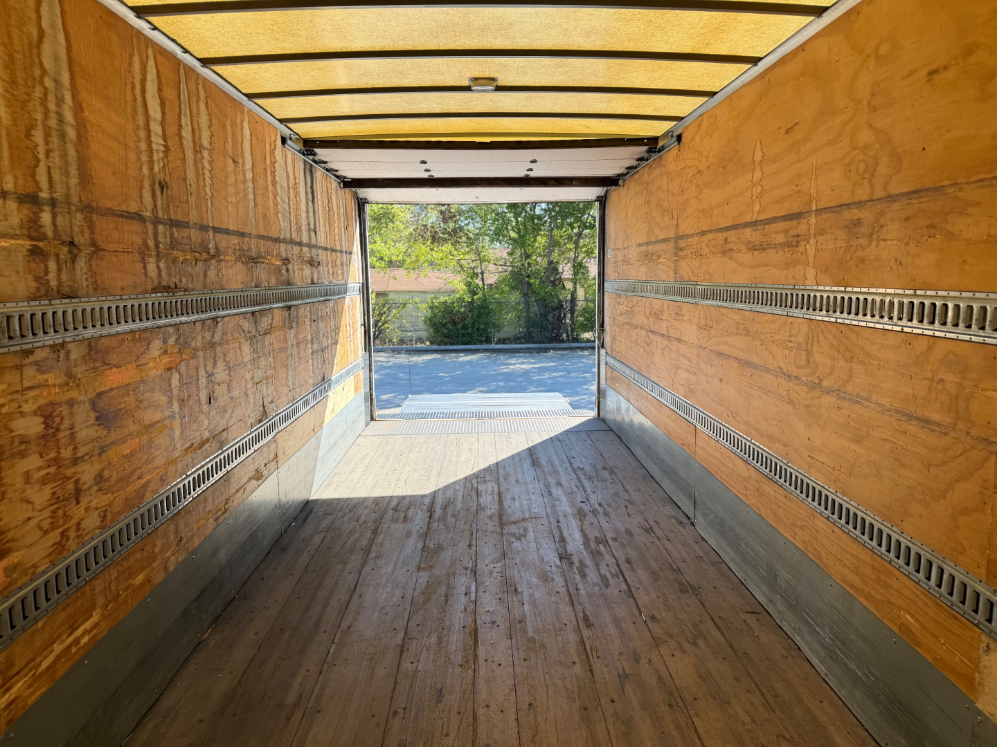 Trailer interior with wooden side walls and metal rails, viewed from open rear door toward a tree-lined outdoor area.