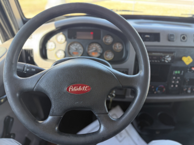 Peterbilt steering wheel and dashboard inside cab. Instrument cluster visible with gauges; center console with controls and small display. Steering wheel badge reads Peterbilt.