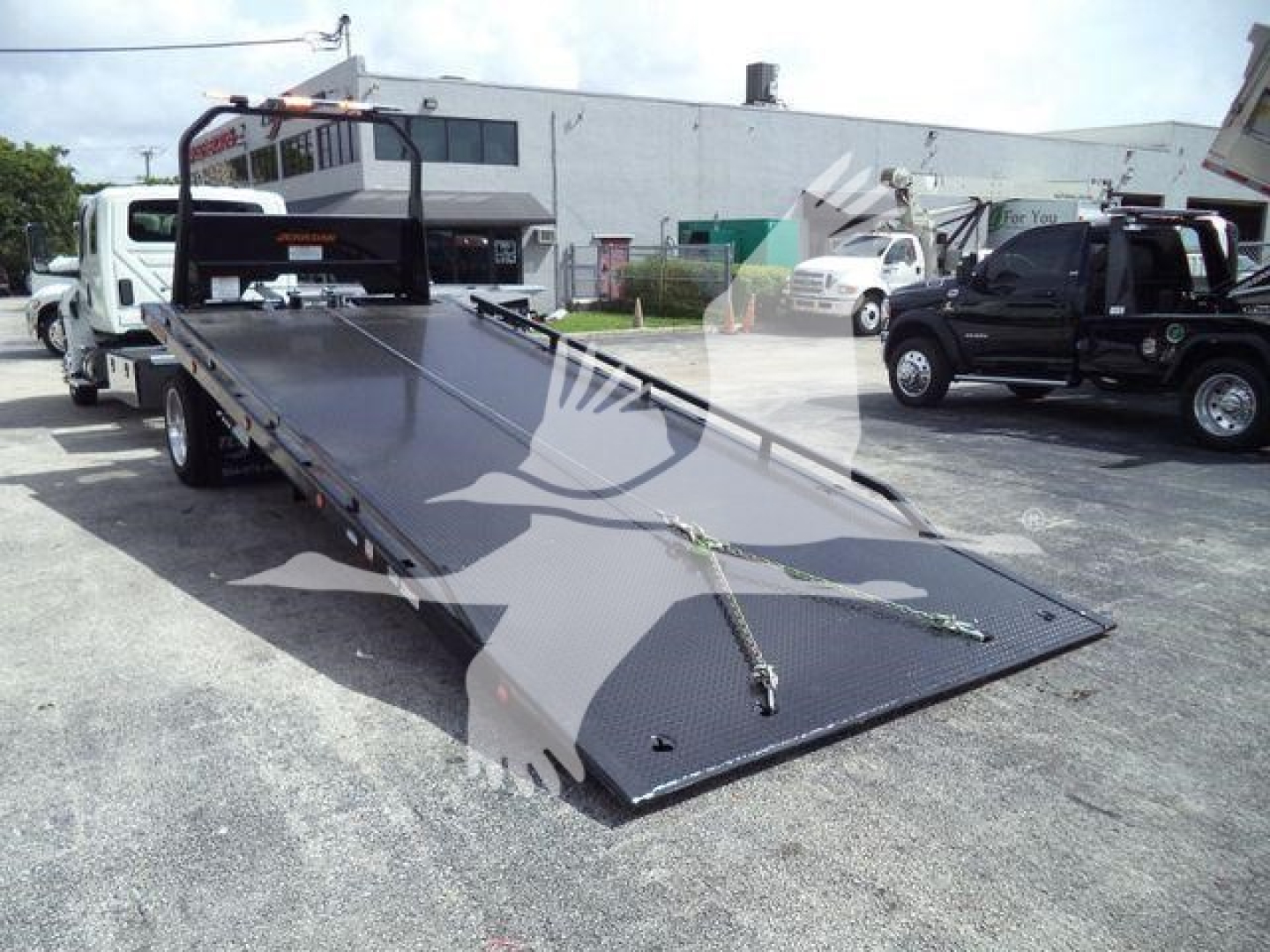 Flatbed tow truck carrier with ramp down, platform surface and safety chains, International MV in background, dealership lot.