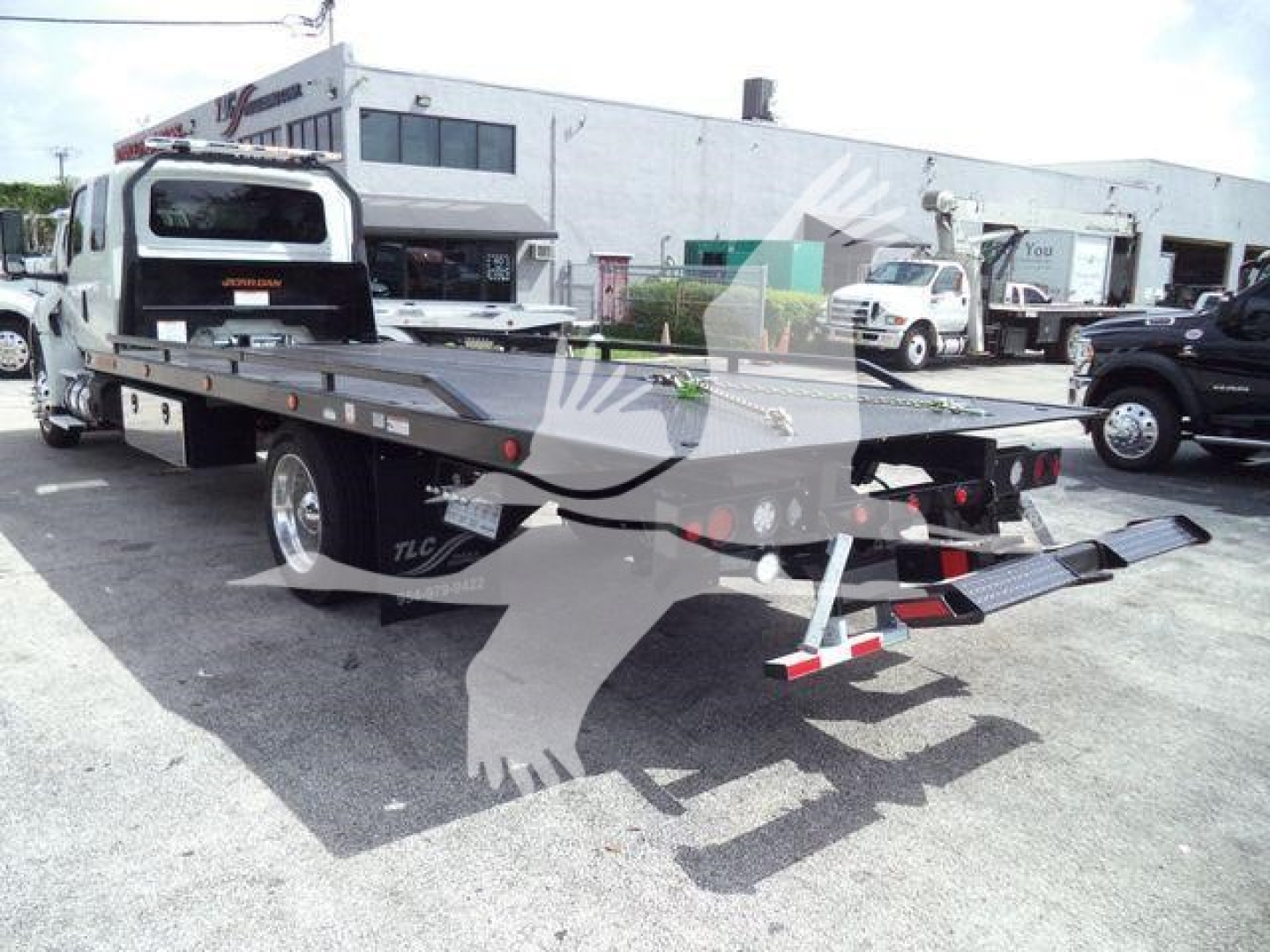 Flatbed rollback truck in a dealership lot, rear view showing loading ramps and hydraulic equipment.