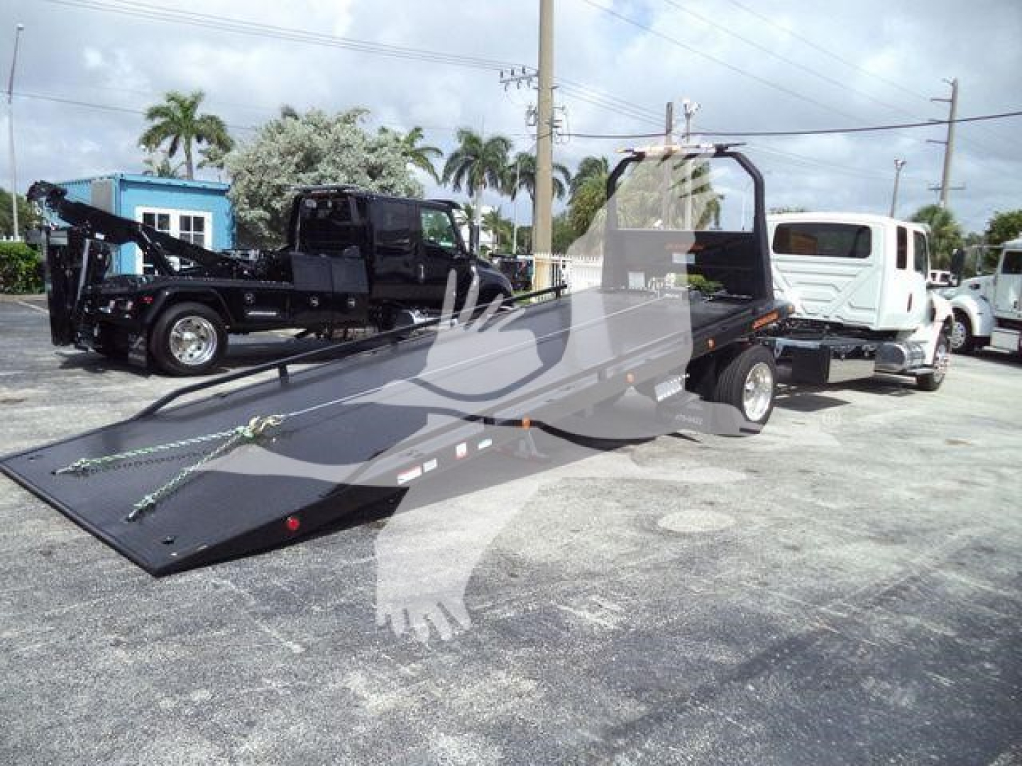 A flatbed tow truck with a ramp on a dealership lot, showing a white cab and a long black flatbed with hydraulic system, other trucks in background.