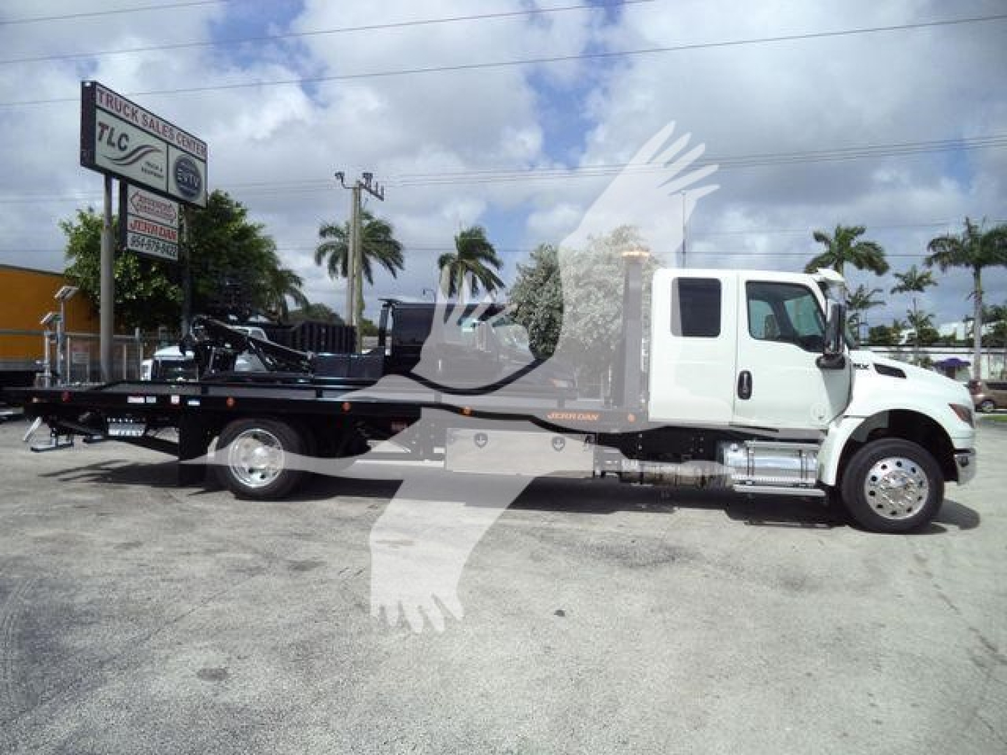 Truck with a flatbed rollback body, white cab, long wheelbase, mounted heavy-duty tow equipment, side steps, and black chassis in a dealership lot.