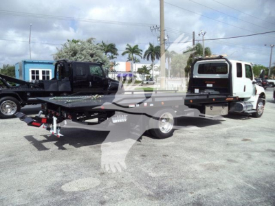 Flatbed rollback tow truck with a white cab and black flatbed, visible equipment and parking lot in background.