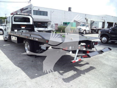 Flatbed rollback truck in a dealership lot, rear view showing loading ramps and hydraulic equipment.