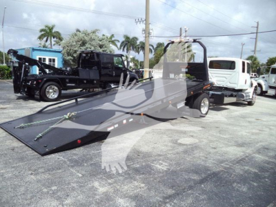 A flatbed tow truck with a ramp on a dealership lot, showing a white cab and a long black flatbed with hydraulic system, other trucks in background.