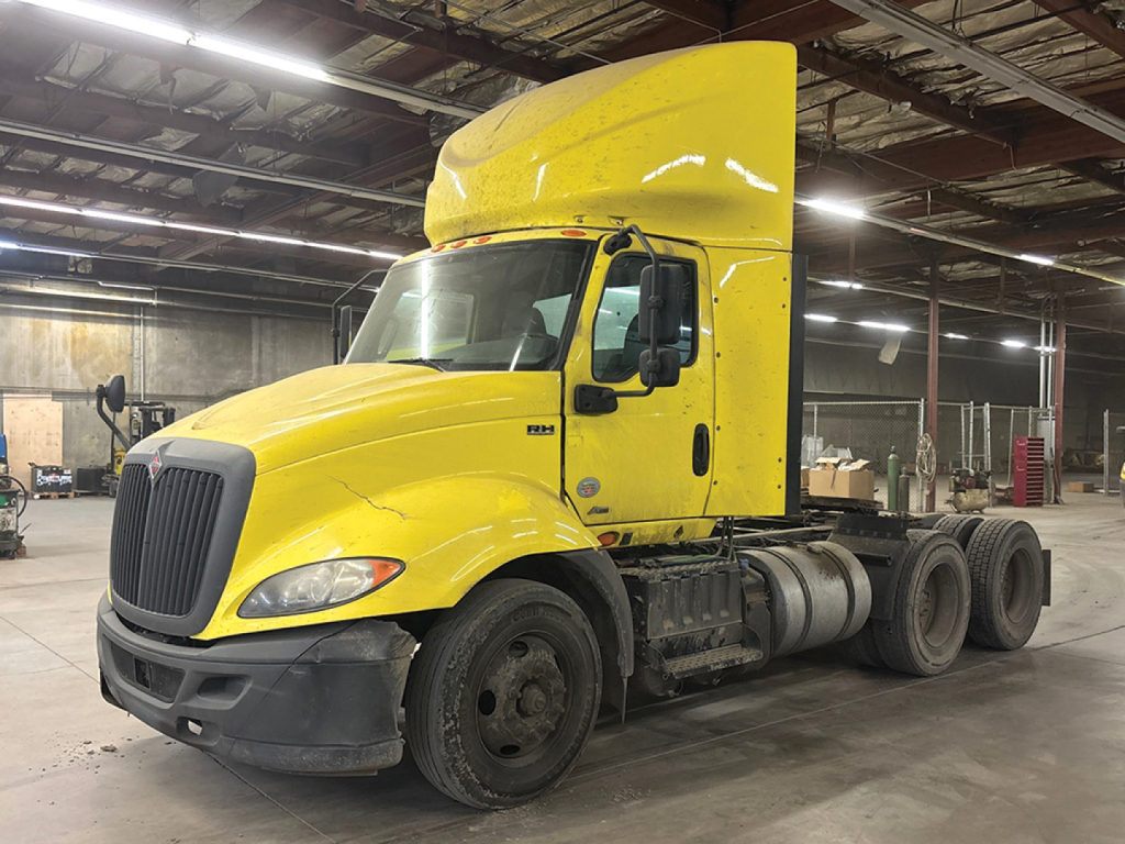 Truck cab and chassis in a warehouse, bright yellow International RH613 daycab, semi-truck with sleeper pod, front grille, and exposed wheels.