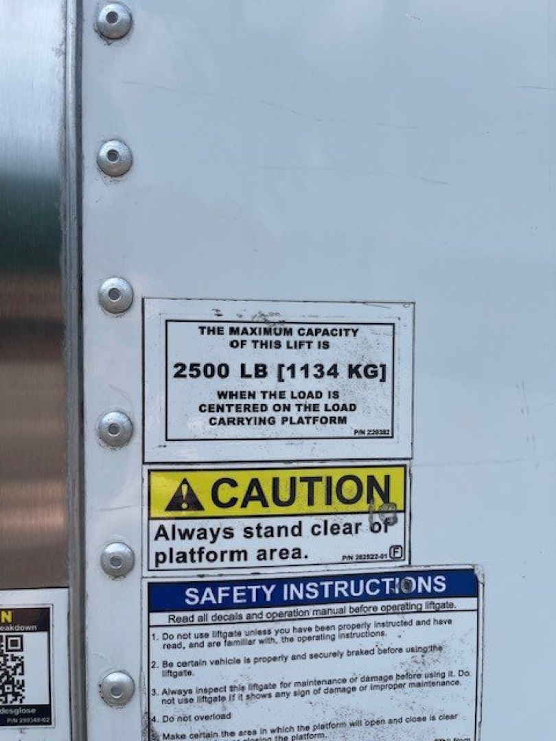 Isuzu NQR truck lift gate maximum capacity plate reading 2500 LB (1134 KG) centered load on platform, caution and safety instructions placards visible on silver lift with bolts.