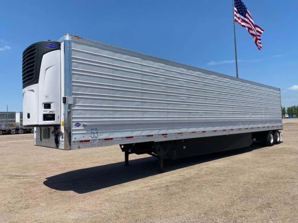 Truck trailer side view showing a refrigerated trailer with a blue-white Carrier/Cooler unit at the front, aluminum ribbed walls, tandem axles, and American flag in background.