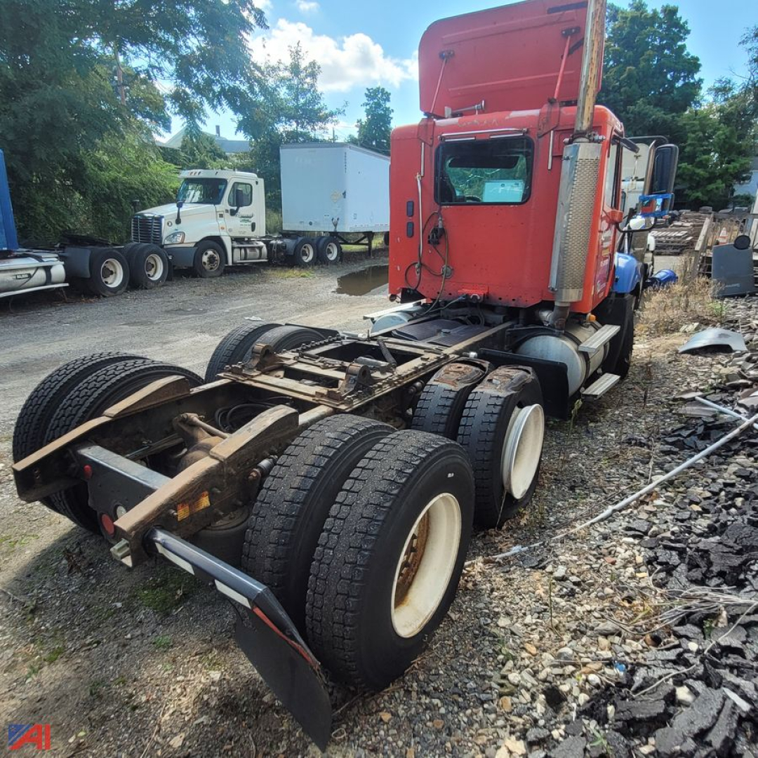 Red Freightliner Columbia 120 tractor truck chassis without cab, rear view showing dual rear axles with white wheels, flatbed frame, and surrounding yard with other trucks.