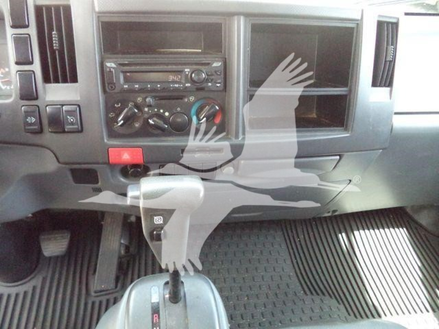 Isuzu NPR-XD dash area showing gear shifter, center console, climate controls, radio, and empty passenger door panel; grey interior with rubber floor mats.