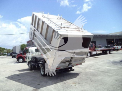 Isuzu NPR XD dump truck in front yard with raised dump bed and various trucks in background.