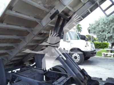 Isuzu NPR truck under a dump body with hydraulic lift mechanism visible and a white truck in background.