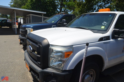 White Ford F-250 Super Duty pickup truck in foreground with orange beacon on roof, blue truck behind, dealership lot setting.