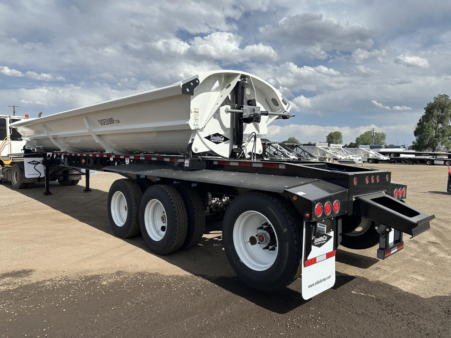 Side-dump trailer showing large white dump body and dual rear axle with tires, rear mudflaps, and hydraulic assembly in side view.