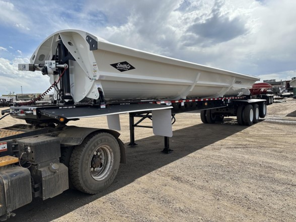 Tri-axle air ride side-dump trailer by Smithco on flatbed truck, white hopper with black Smithco logo, hydraulic mechanism at front, long flatbed with dual rear wheels.