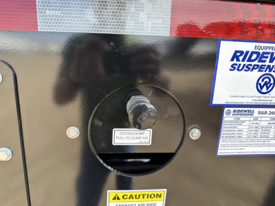 Circular access port on a black trailer body with a label reading 'SYSTEM DUMP PULL TO DUMP AIR' and a Safety sticker. Partial RideRite suspension label visible.
