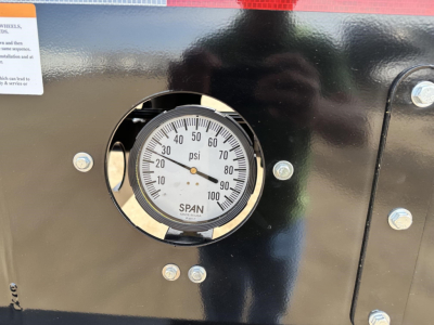 Dial gauge panel showing 0-100 psi scale mounted on black trailer body with metal screws and reflective surface.