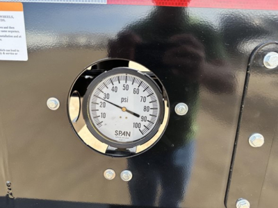 Gauge displaying pressure in psi mounted on black trailer body with bolts around and reflection of person in glossy surface.