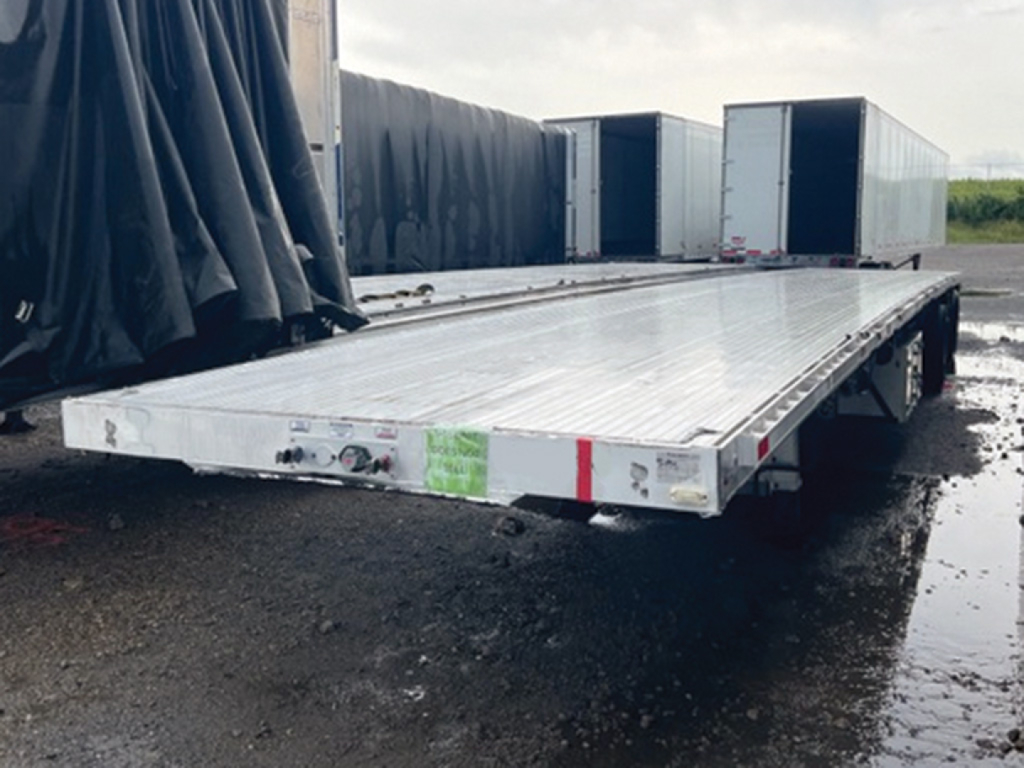 Flatbed trailer with aluminum deck and dual rear ramps, showing long silver platform and storage boxes at rear, outdoor yard setting.