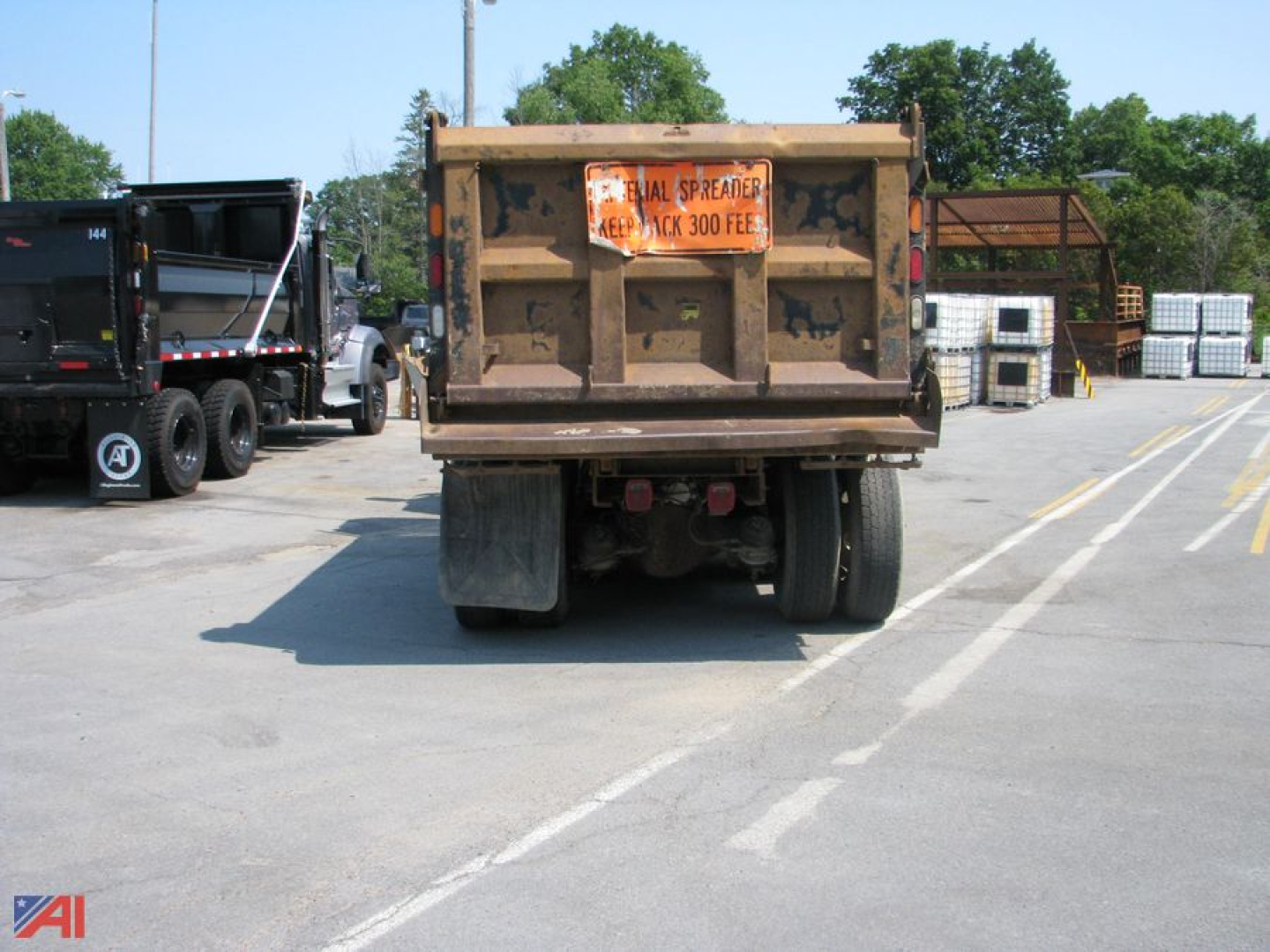 Dump truck rear view showing labeled warning sticker on tailgate and hydraulic components; '1/4 IN. SOLID TIRES' text partially visible on orange plate; industrial yard in background.