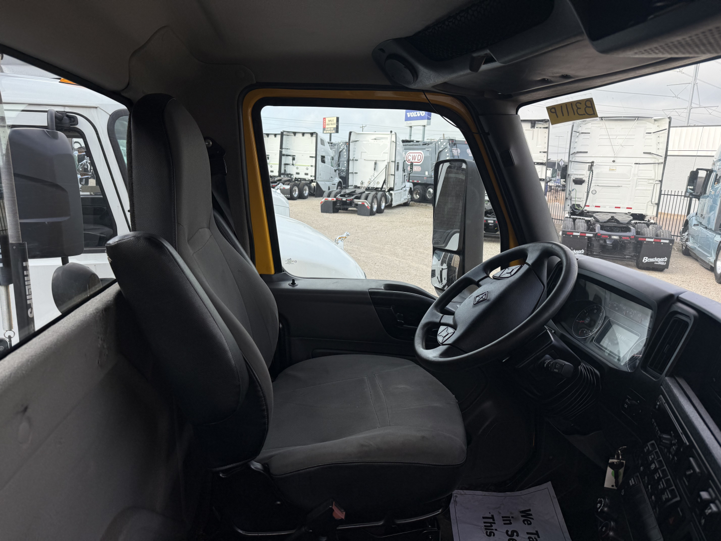 Interior cab of a 2019 International LT truck showing the driver's seat, steering wheel, dashboard, and passenger seat. Visible through the windshield are other trucks and trailers in a dealership lot.