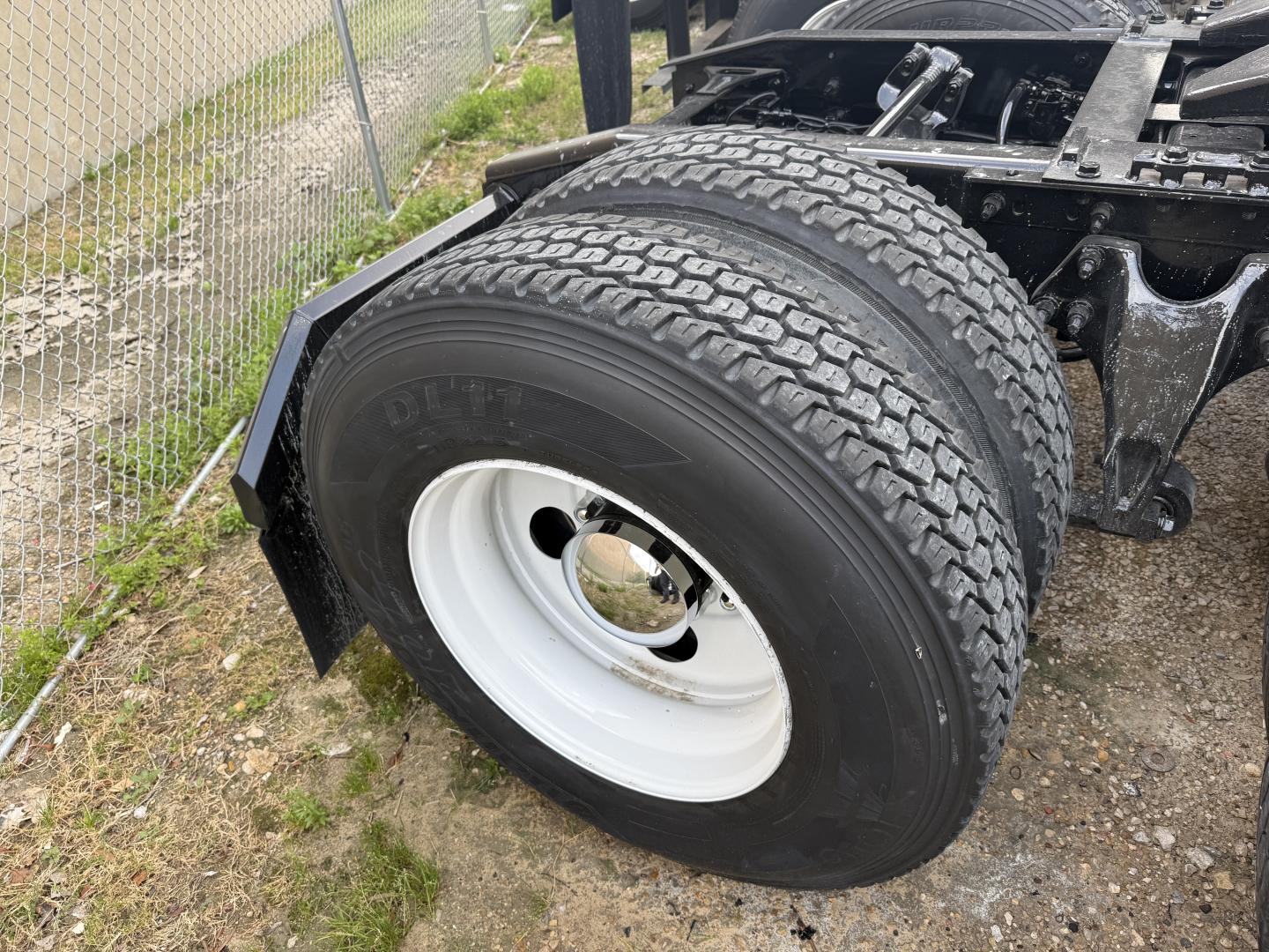 Dual rear wheels of a tractor-trailer chassis showing large black tires and white steel rims; portion of the frame and fender visible.