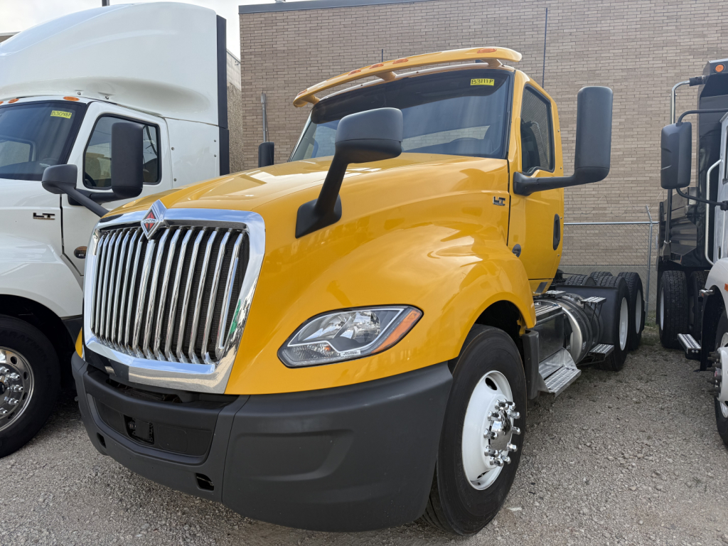 Yellow 2019 International LT conventional daycab tractor showing front grille and mirrors, with other trucks in background.