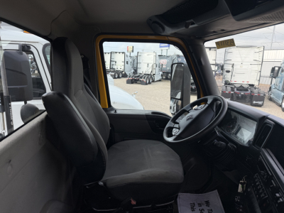 Interior cab of a 2019 International LT truck showing the driver's seat, steering wheel, dashboard, and passenger seat. Visible through the windshield are other trucks and trailers in a dealership lot.