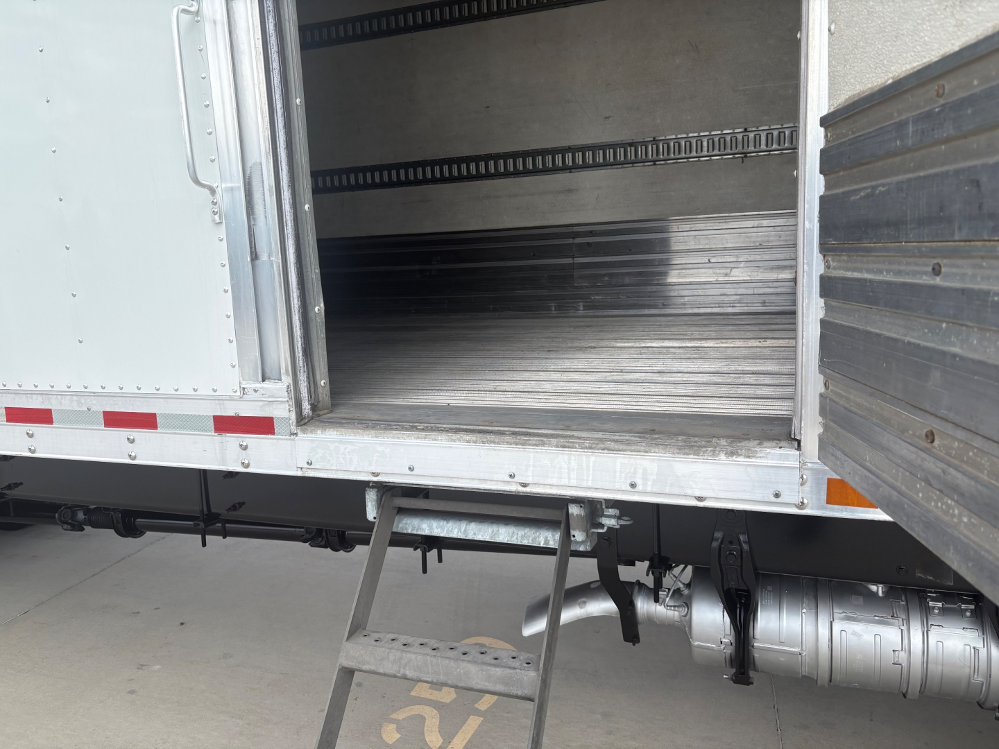 Truck refrigerated trailer interior with metal flooring and side rails; bottom step ladder at cargo door; cylindrical fuel or air tank and drive line visible