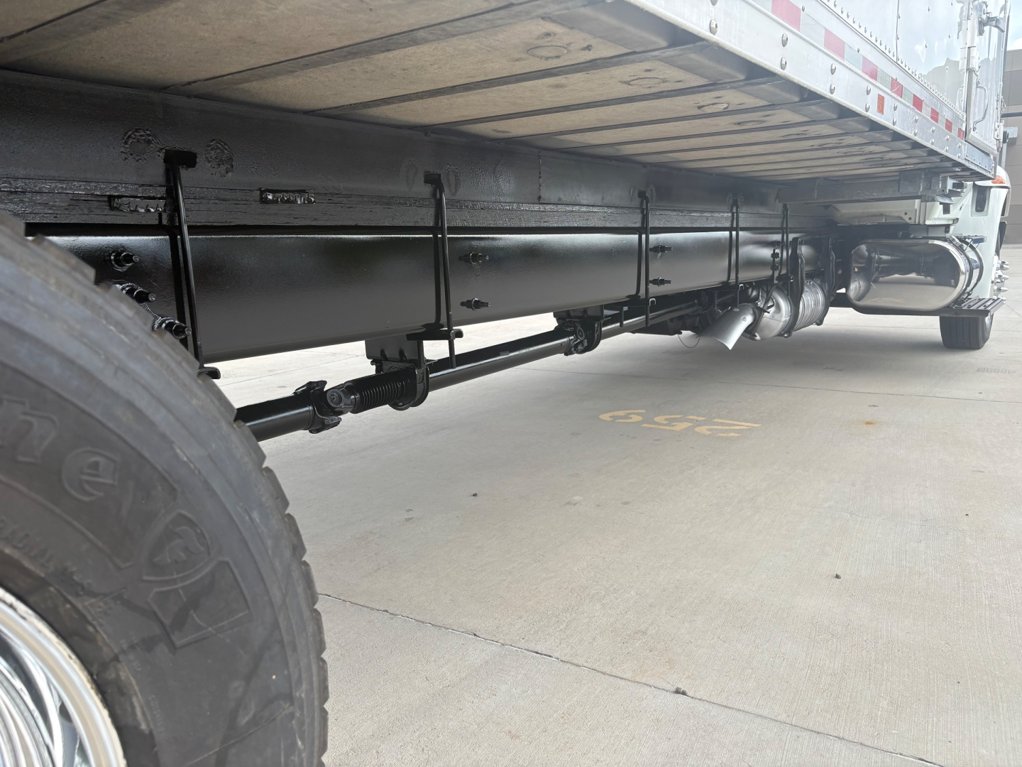 Underbody of a refrigerated truck showing the frame, fuel lines, and a cylindrical component along the side. Visible items include a large tire on the left, a black cylindrical tank or accumulator running parallel to the frame, and piping u