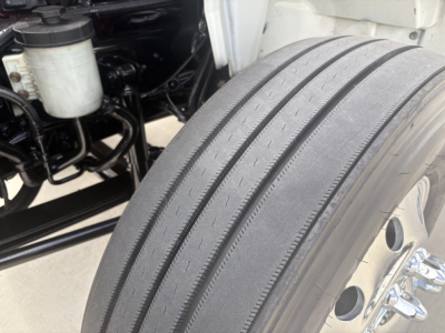 Truck wheel and front suspension with large rear dual steering tires, engine bay visible in background; shows tire tread and wheel well on International MV607 refrigeration truck.