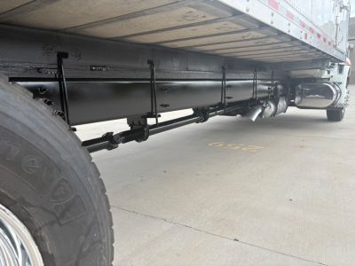 Underbody of a refrigerated truck showing the frame, fuel lines, and a cylindrical component along the side. Visible items include a large tire on the left, a black cylindrical tank or accumulator running parallel to the frame, and piping u