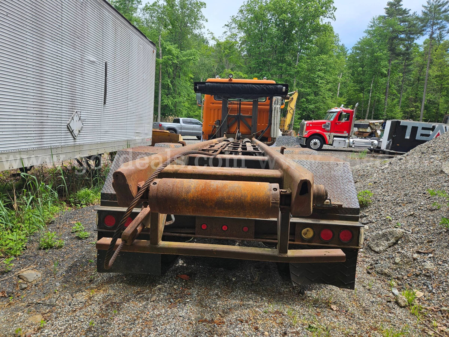 Rear view of a rusty flatbed roll-off truck chassis with a large hook roller and taillight section, outdoor lot with trees and other trucks.