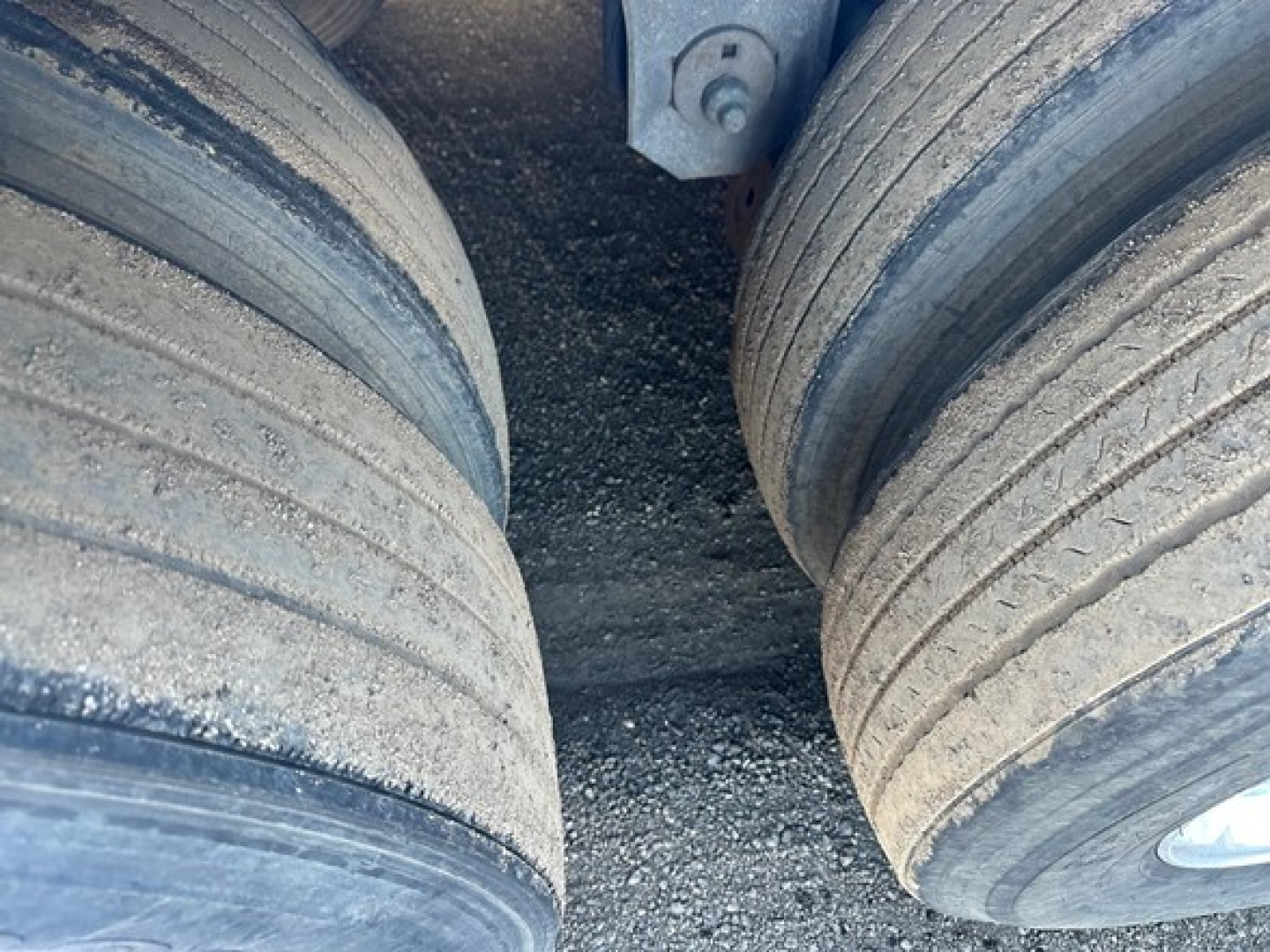 Dual rear trailer wheels with worn tires and gravel surface between them, undercarriage visible.