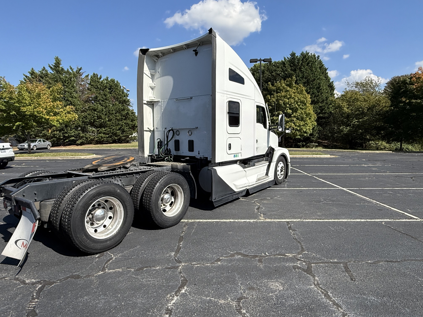 Kenworth T680 tractor in a parking lot viewed from rear-left side, white cab, sleeper, chrome wheels, dual rear axles, sky with clouds.
