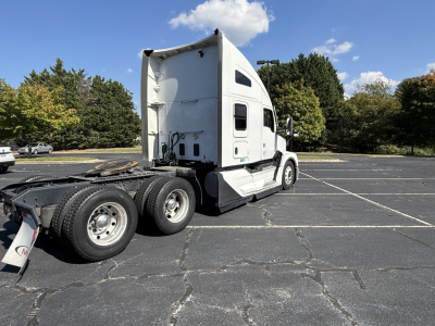 Kenworth T680 tractor in a parking lot viewed from rear-left side, white cab, sleeper, chrome wheels, dual rear axles, sky with clouds.