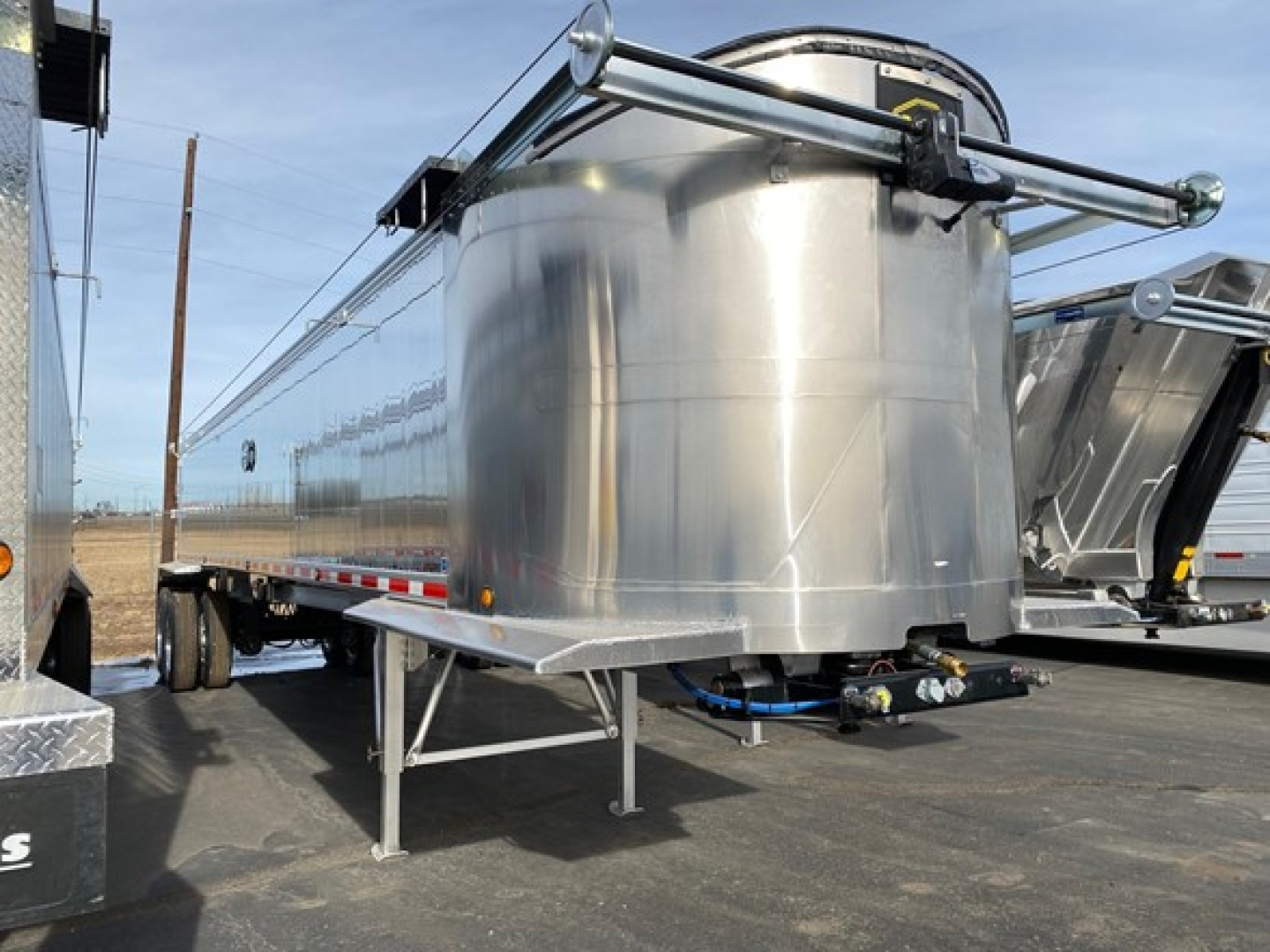 Dump trailer side view with cylindrical stainless steel tank and frame, long flatbed with support legs, hoses and valves visible beneath, chrome railing on top.