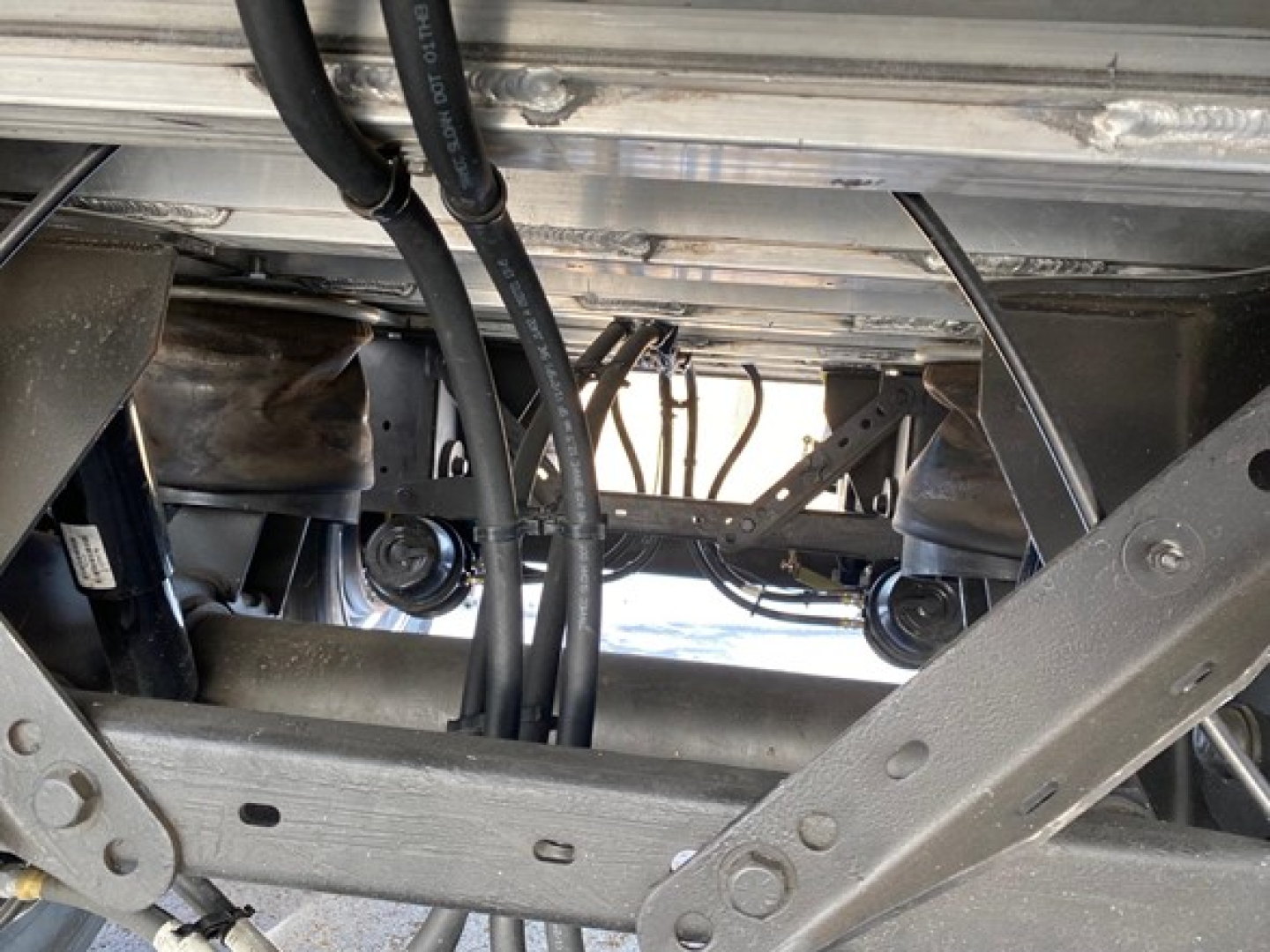 Trailer undercarriage showing steel frame members and hydraulic hoses with wheels visible in background.