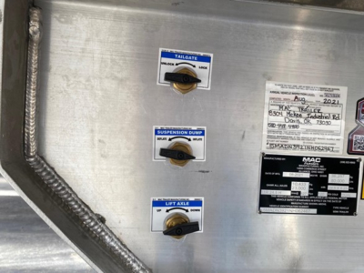 Trailer side panel with three gauges/controls: top labeled TAILGATE with UP/DOWN position indicator, middle labeled SUSPENSION DUMP with UP/DOWN, bottom labeled LIFT AXEL with UP/DOWN; also a metal plate with VIN and other specs, and braide