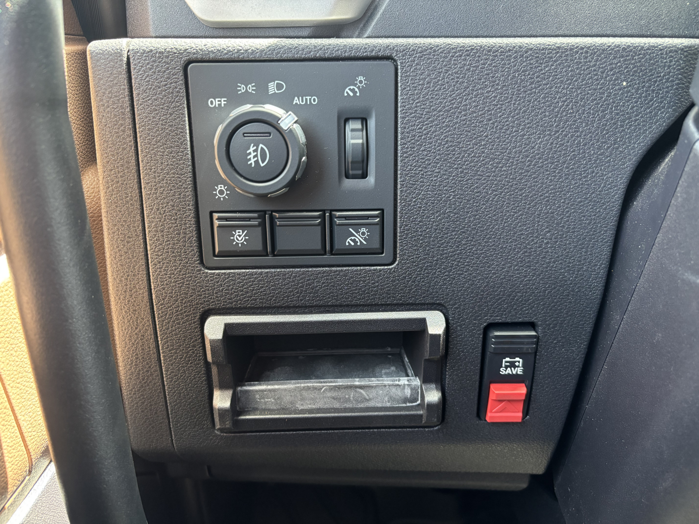Dashboard control panel with round headlight switch and a row of buttons, a small storage slot below, and a red e-stop button labeled 'SAVE' on the right side.