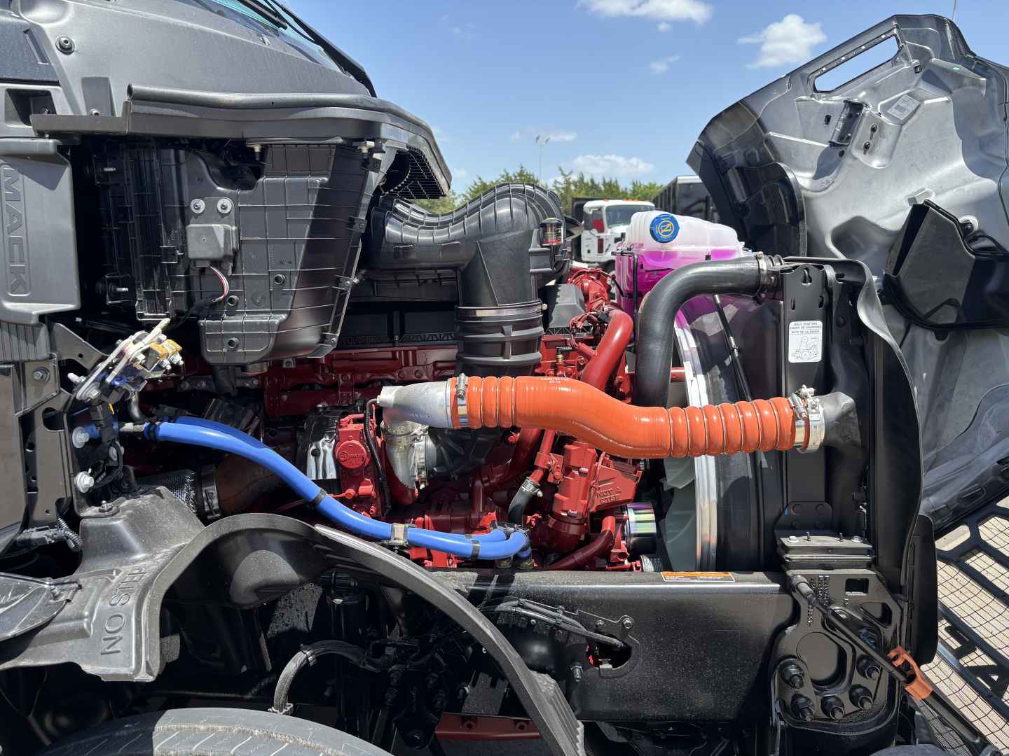 Truck engine bay showing Mack PR64T76H engine with red engine block, blue hose, orange coolant hose, black plastic intake, and purple coolant reservoir cap.