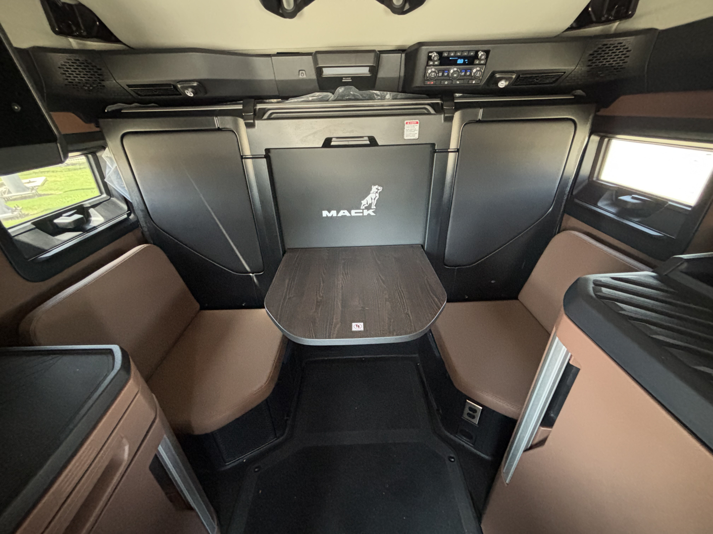Interior of a new Mack PR64T76H sleeper cab showing table, seating, and overhead dash. Visible text includes MACK logo on the partition wall and a control panel above.