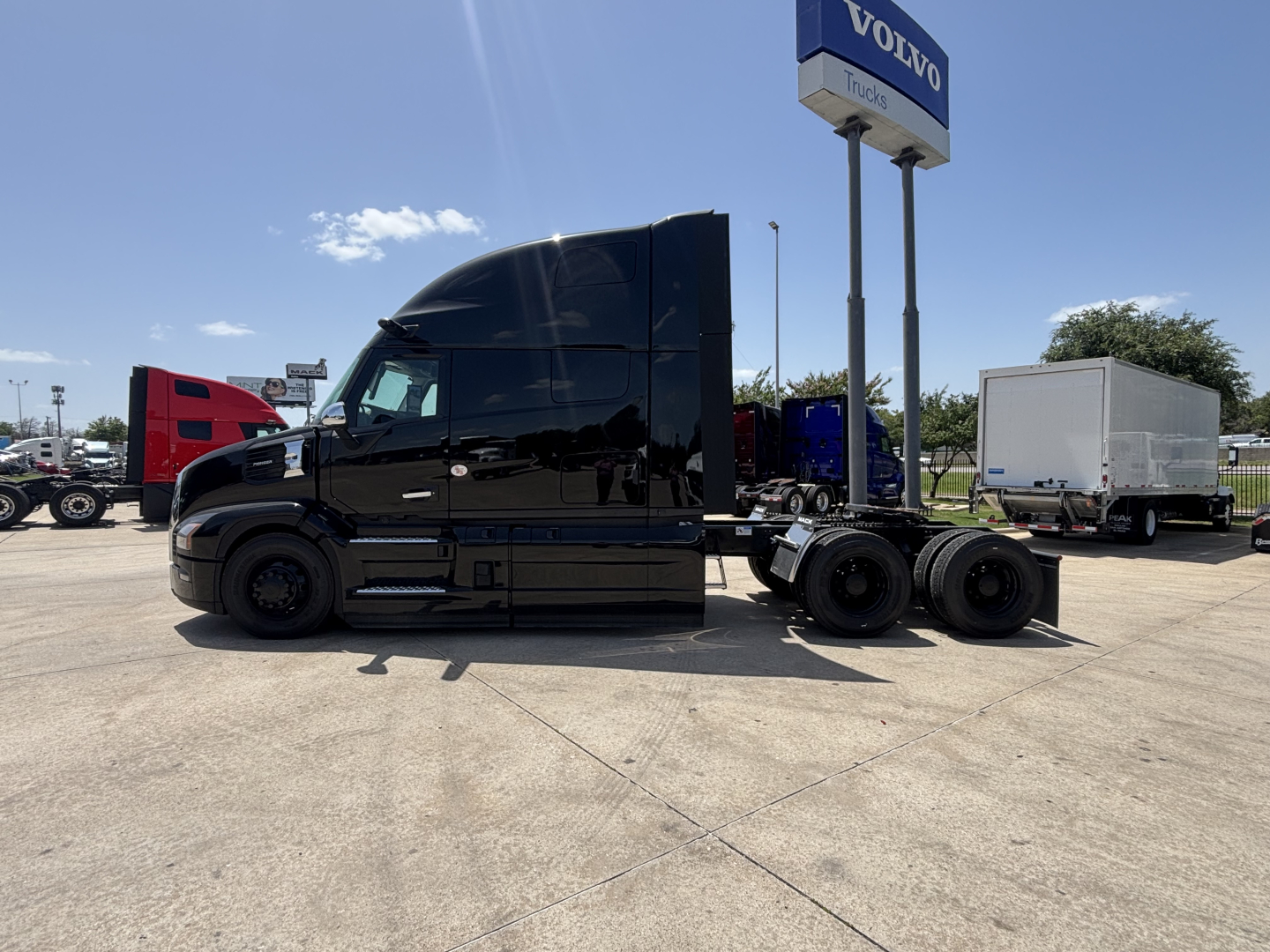 Truck tractor in black, side view showing sleeper cab and rear axles with dual wheels; dealership lot in background with tall Volvo sign.