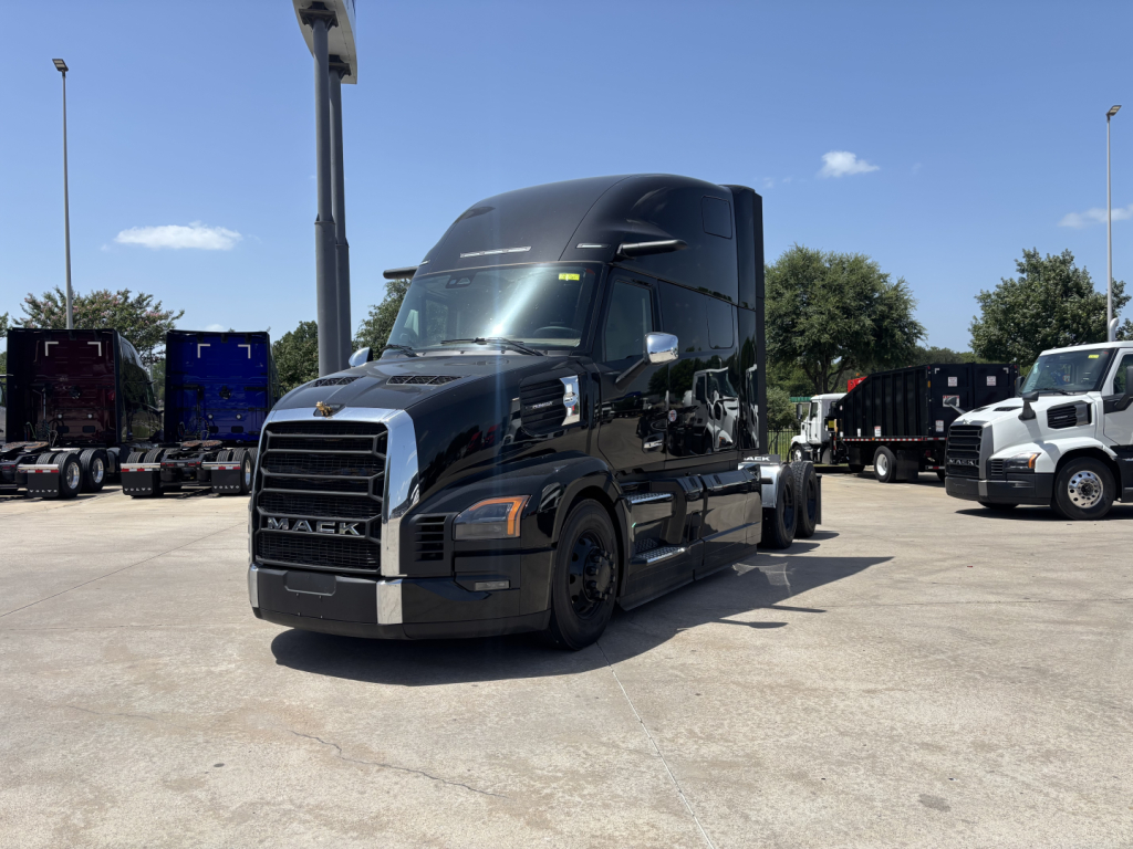 Mack PR64T76H conventional sleeper truck in lot, black exterior, front grille visible, other trucks in background.