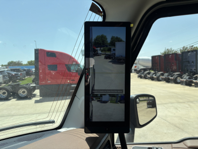Truck cab interior with a vertical tablet display mounted in the window. Exterior view shows a lot with red truck cabins and trailers.