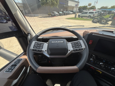 Mack PR64T76H truck cabin steering wheel and dashboard interior with controls on both sides; windshield and outside trucks visible.
