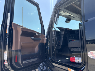 Cab interior of a new 2027 Mack PR64T76H truck with door open and cockpit visible, steering wheel and dashboard seen, brown door panel, seat, and floor mats.